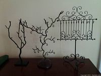 Wide view showing all three metal jewelry stands on a table: two tree-shaped stands and one ornate rectangular stand with hooks.