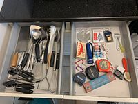Drawer with large kitchen utensils including spoons, ladle, can openers, whisk, grater, measuring tapes, and small tools.