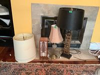 Three assorted lamps with shades including a plaster base lamp with cream shade, a metal and glass base lamp with pink shade, and a wood lamp replica with black shade.