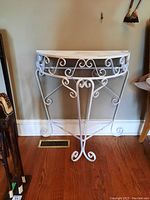 White painted metal half moon table placed against a wall on hardwood flooring. Shows decorative scroll designs on legs and edges with a flat top surface that has discoloration and stains.