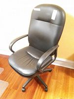 Black office chair with cushioned seat, backrest, armrests, and caster wheels, placed on wooden floor against yellow wall.