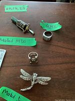 Four jewelry items on a wooden surface with green labels indicating markings: a band ring marked 925, a ring marked MTA with round stone, a stone pendant, and a dragonfly brooch with rhinestones