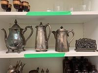 Three silver-plated pots (two coffee pots and a teapot) and a metal napkin holder displayed on a shelf, showing tarnish on all items.