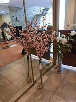 Two tall glass vases with faux floral stems, standing on floor in front of a mirrored wall, showing the flowers and rocks inside each vase.