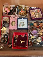 Wide overhead shot showing multiple small boxes and trays of assorted costume jewelry including pins, earrings, and bracelets arranged on a wooden surface.