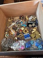 Top-down view of a drawer containing various costume pins and earrings including beads in plastic bags at the bottom, assorted gold-tone and colorful pins and brooches above.