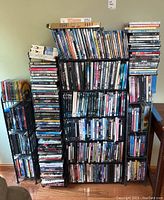 Full view of multiple black metal DVD stands filled with DVDs in cases, stacked and arranged on hardwood floor against light wall