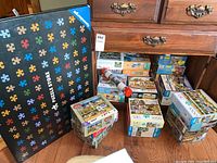 Overall view of various boxed jigsaw puzzles stacked on the floor near a wooden dresser. Large black puzzle store box with multicolored puzzle piece graphics also visible.