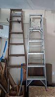 Wooden and aluminum 6-foot ladders standing side by side against a garage wall.