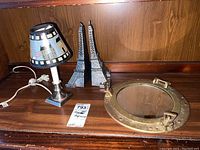 Photo showing table lamp with a navy lampshade with nautical design, metal Eiffel Tower bookends, and round brass ship's porthole mirror on wooden surface