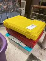 Stack of three plastic cases: yellow Construx carrying case on top, followed by red and clear blue lidded containers below.