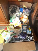 Box of assorted untested light bulbs including boxed packs and loose bulbs in various packaging, placed on the floor between wooden furniture.