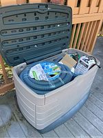 Wide shot showing beige and gray plastic outdoor storage chest outdoors with lid open containing pool accessories including a coiled blue and black vacuum hose.