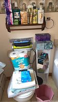 Photo of bathroom shelf with multiple toiletries including hair care, mouthwash, soaps, pads, tissue boxes and large pack of toilet paper on toilet tank, also razors and personal care items on stand.