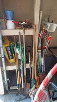 Wide view of yard tools leaning against a cabinet and shelf in a garage closet area showing various shovels, rakes, pruners, and lawn care products.