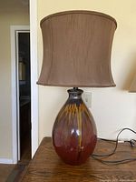 Photo shows a brown ceramic table lamp with a rounded glossy base with multiple brown hues and a dark brown silk lampshade on top. The lamp is placed on a wooden table.