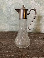 Wine decanter with cut glass body and silver-colored metal top and handle shown in front of textured background.