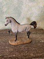 Horse figurine with raised front leg on base, light gray with darker accents, signed and numbered by artist