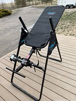 Photo of a black inversion table on a wooden deck outdoors showing the metal frame, padded backrest, ankle holders with foam pads, and hand grips.