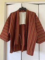 Front view of red cotton Komono jacket hanging on black hanger. Jacket has multicolored vertical stripes in red, brown, and beige and wide sleeves. Open front with tie closure and white lining at upper back.