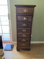 Front view of a tall, slender lingerie chest made of solid maple with six drawers and brass handles, placed on a wood floor near a sliding door.