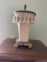 Old fashioned night light with cream cloth covering, tasseled trim, and wooden base on table.