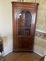 Solid cherry wood corner china cabinet with glass door divided by wooden muntins and lower wooden storage door, positioned in corner of room next to floral wallpaper border.