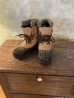 Pair of brown and tan Sorel boots placed on wood surface, showing front and side profiles; boots feature waterproof rubber lower and warm lining.