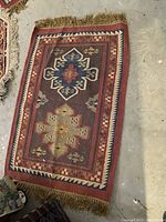 Photo of a small wool rug with geometric patterns in earthy colors and fringes on the edges, placed on a concrete floor.