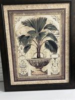 Photo of framed botanical plant art print showing a green palm plant in an ornate urn on a vintage style patterned background with a dark wood frame