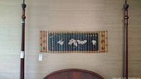 Horizontal image showing the painting mounted on the wall between two wooden bedposts. The painting has visible wood slats, a dark green background with white floral figures and golden ornamental border.