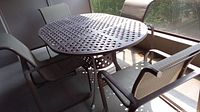 Four metal framed patio chairs around a round table on a screened porch with greenery outside. Chairs have brown weather resistant fabric seats and backs.