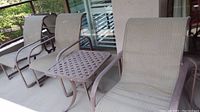 Four patio chairs with metal frames and woven fabric seats and backs in beige color; arranged on outdoor porch.