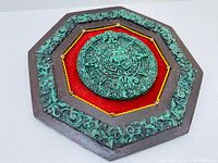 Front view showing octagonal wooden base with cast resin green Aztec calendar medallion on red fabric background.