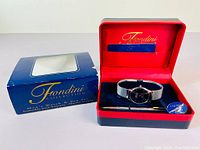 Open red and black clamshell box with red lining, containing the men's wristwatch with black dial and silver mesh band, along with a pen and Fondini branding inside.