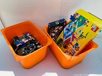 Two orange storage bins filled with assorted LEGO pieces including boxed kits and instruction manuals