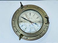 Front view of the brass porthole style ship's clock showing the clock face and brass frame with patina and clasps.