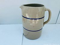 Side view showing the beige stoneware body with two blue horizontal decorative bands and the handle.