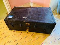 Closed view of the rectangular black metal clad trunk with metal corner reinforcements, rivets, central lock and side latches.