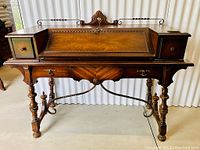 Front view of walnut spinet desk showing carved crest, fold-down writing surface, turned legs and wrought iron stretcher.