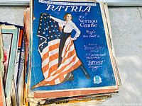 Photo showing top sheet music with blue cover titled 'Patria' featuring a person holding an American flag, illustrating patriotic theme.