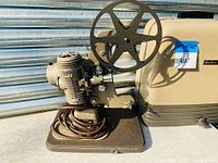 Side view of parkerized metal Bell & Howell projector showing film reel mount and power cord coiled on base.