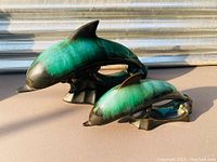 Two ceramic dolphin figurines in green and black glaze positioned side by side, showing their left side profiles and bases.