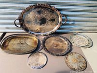 Photo showing six silverplated trays and coasters of various shapes and sizes with detailed engraving and some tarnish, all in very good vintage condition.