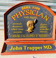 Full view of wooden physician sign with removable nameplate 'John Trapper MD' showing all painted text and carved relief profile.