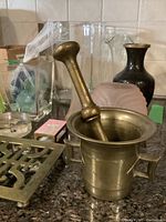 Brass mortar and pestle centered on countertop with other glassware and kitchen items behind.