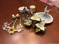 Photo showing assorted porcelain decorative items including floral stems, flower cluster, bird figurines, and small cup from a frontal view on a wood surface.