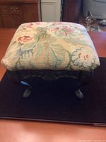 Front view of square footstool showing floral needlepoint cushion and black cast iron base with claw feet on black surface.