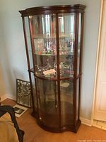 Front angled view of wooden curio cabinet with curved glass doors, illuminated interior, reflective mirrored back and multiple glass shelves. Items inside display excluded from sale.