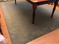 Full view of large green rug laying flat with pattern and part of table legs visible, showing general condition and size relative to furniture.